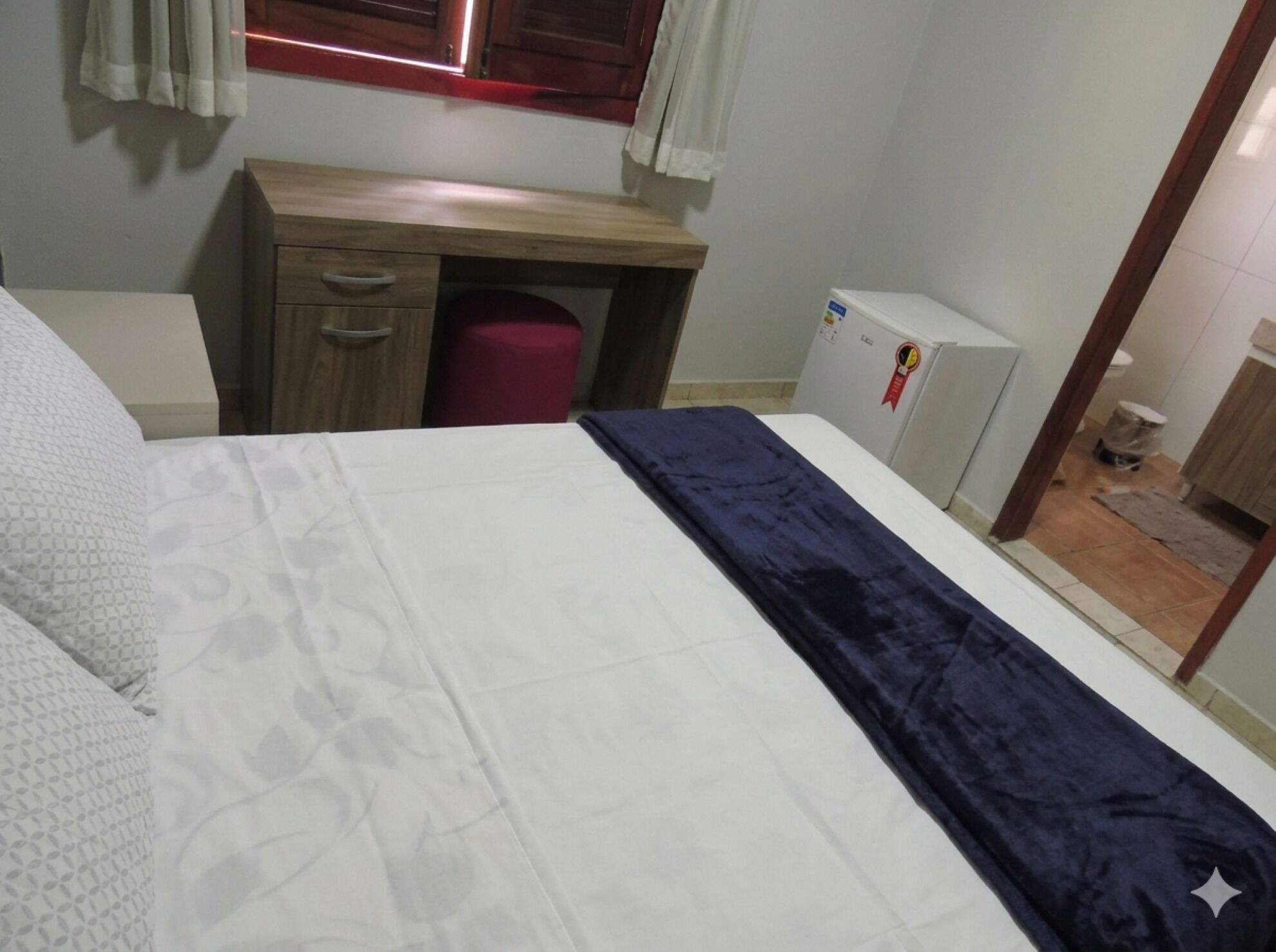A bedroom with a bed, a wooden desk, a red trash bin, and a white refrigerator.