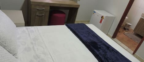 A bedroom with a bed, a wooden desk, a red trash bin, and a white refrigerator.
