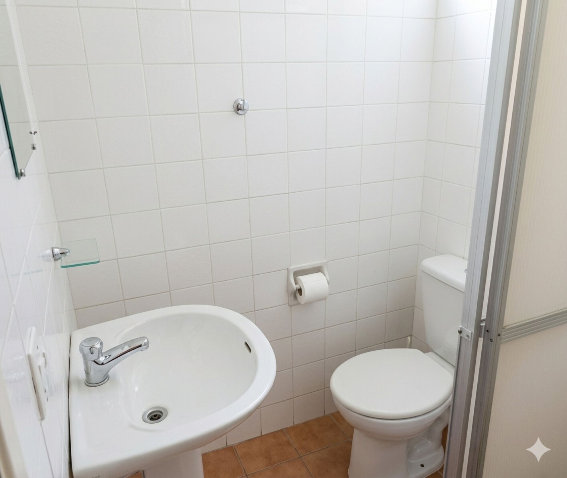 Casa de banho pequena com lavatório branco, sanita e porta de chuveiro em vidro.