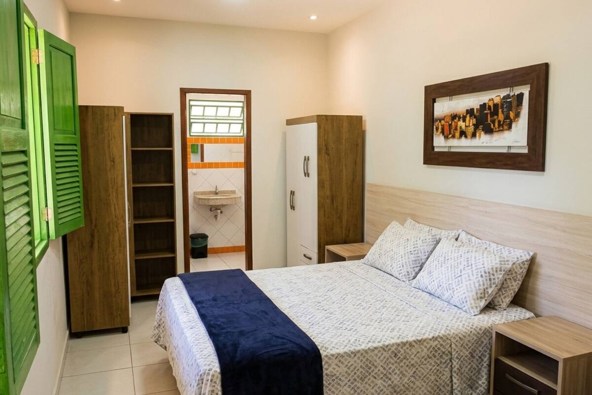 A bedroom with a bed, a wardrobe, a nightstand, a framed picture on the wall, and a bathroom visible through an open door.