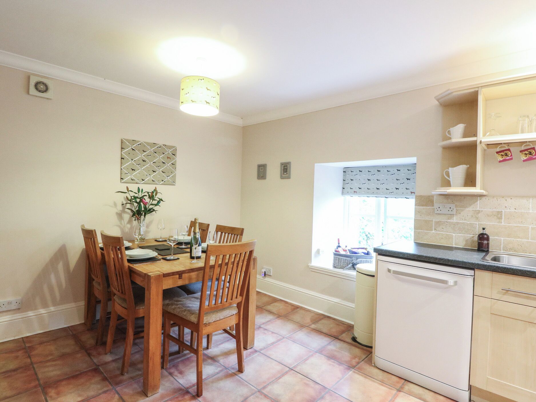 A dining area with a wooden table and chairs, a vase with flowers, and a bottle of champagne. There is a window with a patterned blind and a kitchen area with a dishwasher and cabinets.