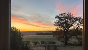 Une vue par la fenêtre sur un coucher de soleil au-dessus d’un champ, avec un arbre et une clôture.