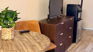 A room with a wooden table, a potted plant, a remote control, a TV on a stand, and a dresser. - The Windmill Motel (Olney)