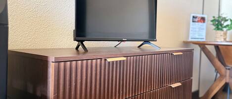 A wooden cabinet with a flat-screen TV on top, a wooden table with a plant, and a framed picture on the wall.