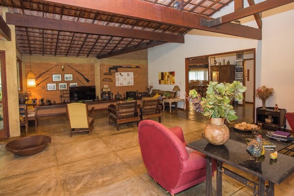 A spacious living area with a high ceiling, wooden beams, and a fireplace. There are multiple seating options including a red armchair and a sofa. A dining table is visible in the background. - Pousada Verbicaro - Santa Maria Madalena (Santa Maria Madalena)