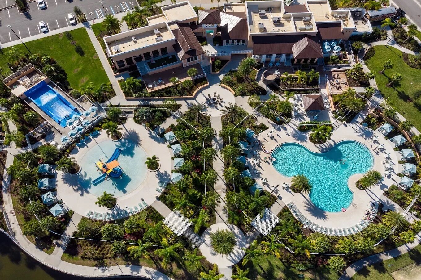 Vista aérea de un resort con dos piscinas grandes, un área de juegos infantiles y diversas zonas de estar al aire libre, todo rodeado de palmeras.