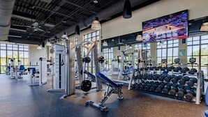 Un gimnasio con diversos equipos de ejercicio, incluyendo máquinas de pesas y mancuernas, y una gran pantalla mostrando un evento deportivo.