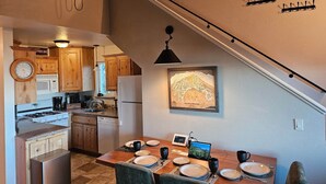 A modern kitchen with wooden cabinets, a dining area with a table set for four, and a staircase with a light fixture.