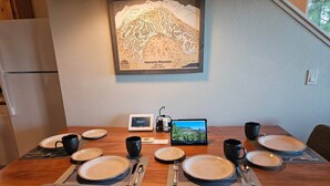 A dining area with a wooden table set for four, a framed map of Heavenly Mountain, and a tablet displaying a scenic view.
