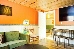 Un espacio de sala acogedor con un sofá verde, sillas de madera, una mesa de comedor y un televisor montado en la pared.