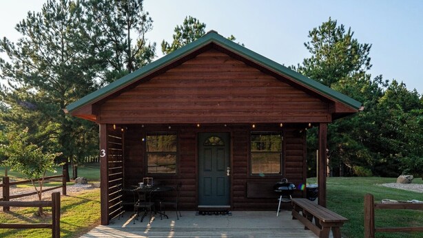 A wooden cabin with a porch, a grill, and a table with chairs. - Timber Lodge Ranch - Studio 3 (Amity)