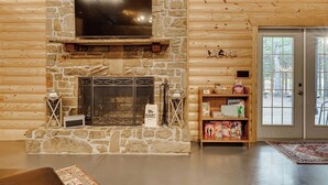 L'intérieur d'une cabane en rondins, avec une cheminée en pierre, un téléviseur à écran plat fixé au mur et une bibliothèque contenant des jeux pour enfants.
