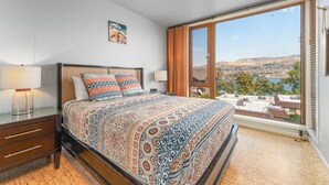 Une chambre à coucher avec un grand lit, une table de chevet, une lampe et une vue sur un paysage, visible à travers une porte-fenêtre coulissante.