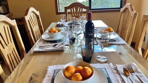 A dining table set with bowls of fruit, wine glasses, and cutlery, with a view of trees through a window. - The BEST VIEW, Rustic Retreat with Hot Tub, Private Fenced Yard, 3BR in Upper Moonridge! (Big Bear City)