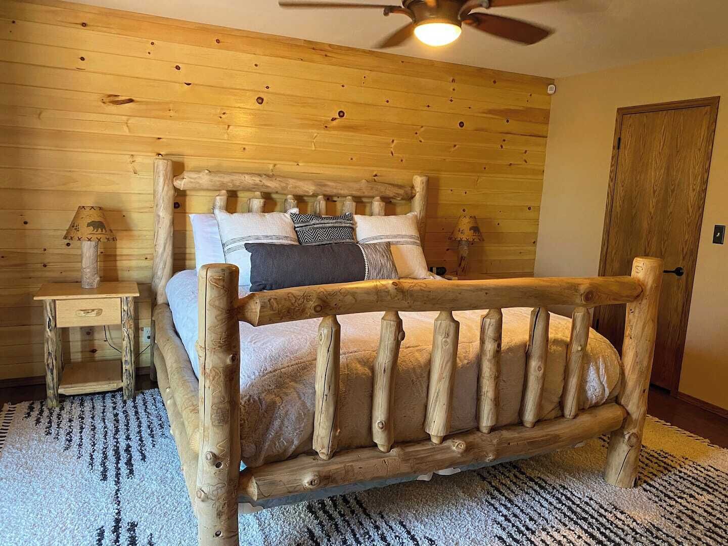 Une chambre à coucher de style chalet, avec un lit en bois, une table de chevet, une lampe et un ventilateur de plafond.