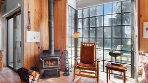 A cozy room with a wood-burning stove, a rocking chair, and a large window offering a view of the outdoors. - Black Butte Ranch Country House 62 (Sisters)