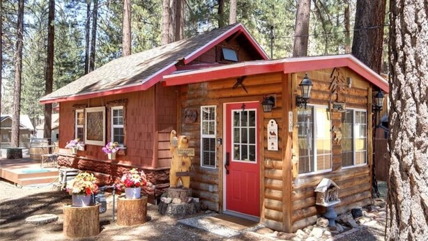 Eine rustikale Holzhütte mit roter Tür, einem Willkommensschild und einer kleinen Veranda mit Blumen.