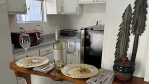 A dining area with a wooden table set for two, white chairs, a wine bottle, and a decorative plant. - Cute, Cozy, Peaceful Getaway, Fenced Yard, Big Bear's Lazy Neighborhood with Soaring Pines (Sugarloaf)