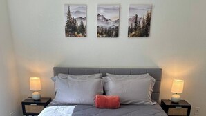 A neatly made bed with a grey quilt, two pillows, a red accent pillow, and a bedside table with a lamp. There are three framed pictures on the wall above the bed. - Cute, Cozy, Peaceful Getaway, Fenced Yard, Big Bear's Lazy Neighborhood with Soaring Pines (Sugarloaf)