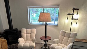 A cozy living room with two rocking chairs, a small table, a lamp, a fireplace, and a window with a view of greenery. - Cute, Cozy, Peaceful Getaway, Fenced Yard, Big Bear's Lazy Neighborhood with Soaring Pines (Sugarloaf)