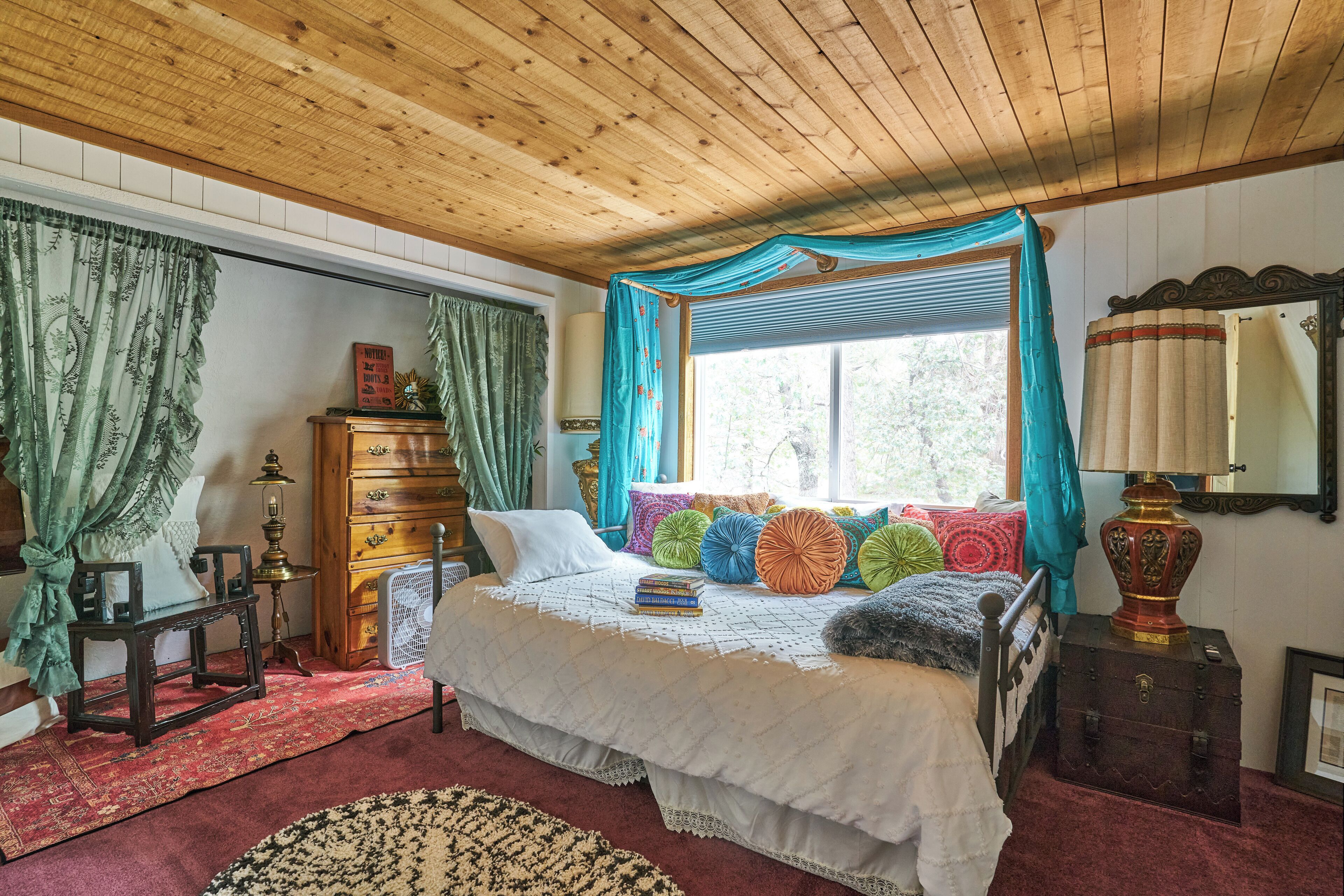 A cozy bedroom with a wooden ceiling, a large bed with colorful pillows, a dresser, and a window with curtains.