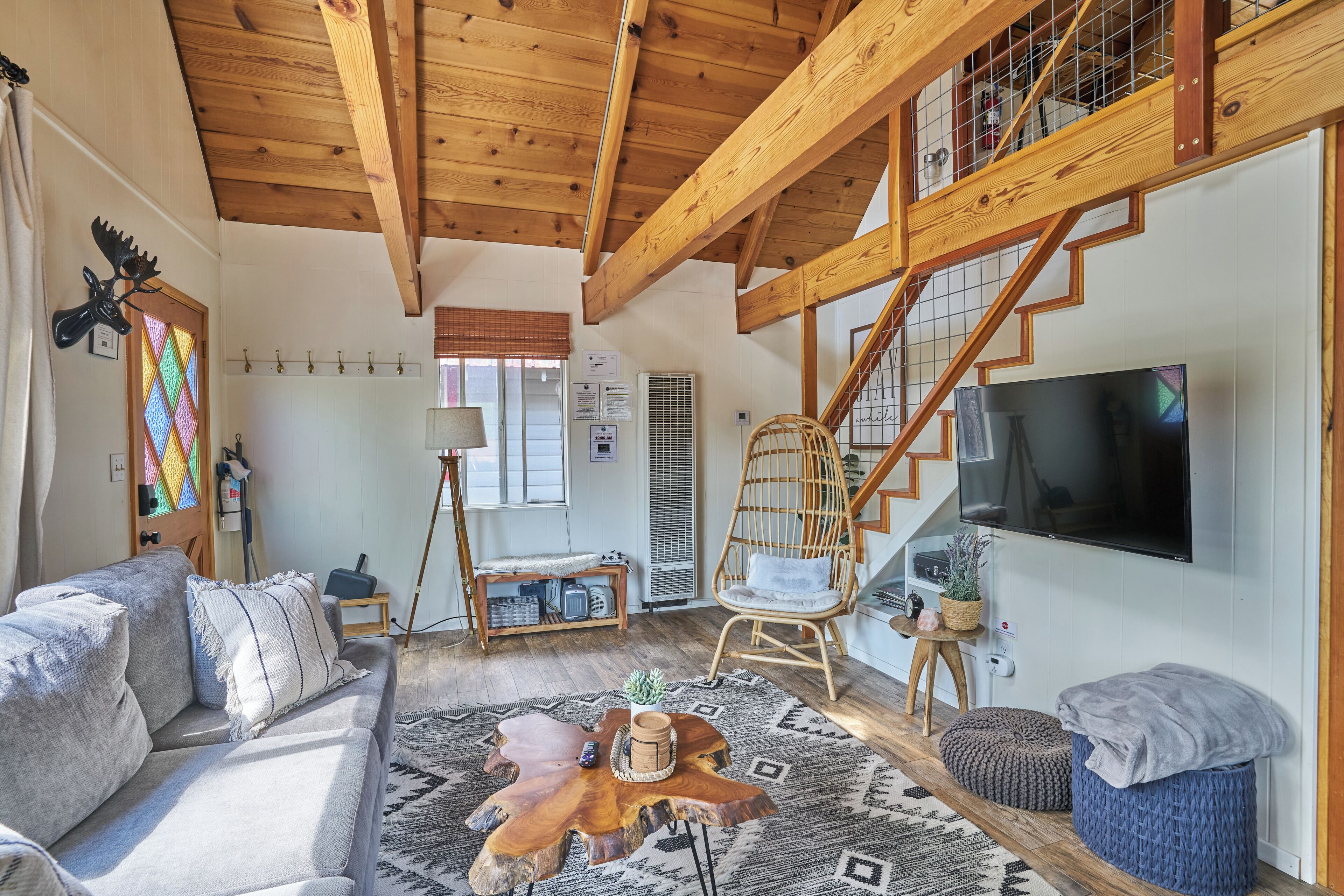 Un salon spacieux avec un plafond haut, des poutres en bois, une grande télévision, un canapé confortable, un fauteuil en osier et un escalier.