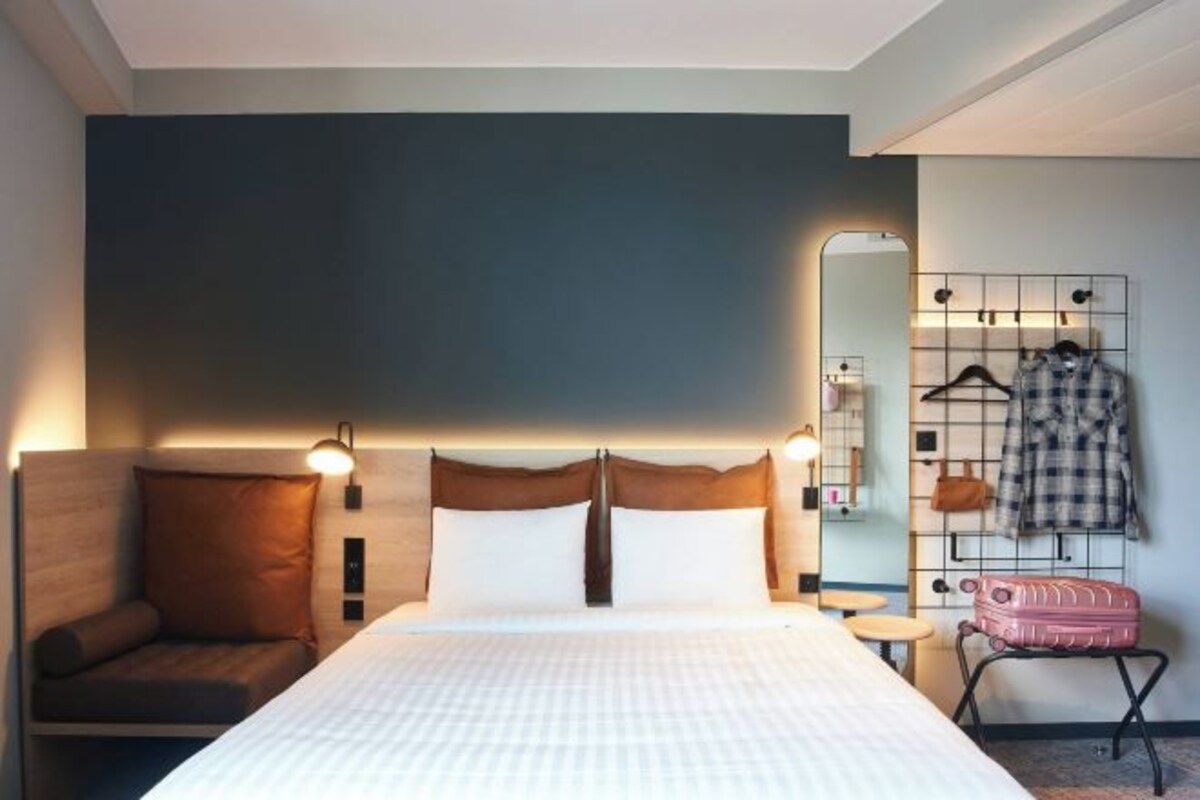 A hotel room with a large bed, a bedside table, a chair, a mirror, and a coat rack with clothes and a suitcase.