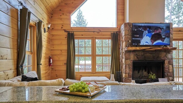 A cozy cabin interior with a stone fireplace, a television mounted above it, and a dining area with a granite countertop and a fruit platter. - Peaceful Rustic Escape, Cool Deck with Firepit Table - No Better Escape than Our Private Lot - 3BR w/Game Loft, Pac Man tabletop games console! (Big Bear)