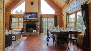 A cozy cabin interior with a stone fireplace, wooden walls, and a dining area with a table set for four. - Peaceful Rustic Escape, Cool Deck with Firepit Table - No Better Escape than Our Private Lot - 3BR w/Game Loft, Pac Man tabletop games console! (Big Bear)