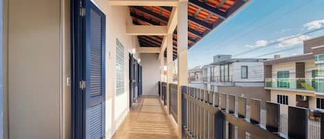 A corridor with a wooden floor, blue shutters, and a view of neighboring buildings.