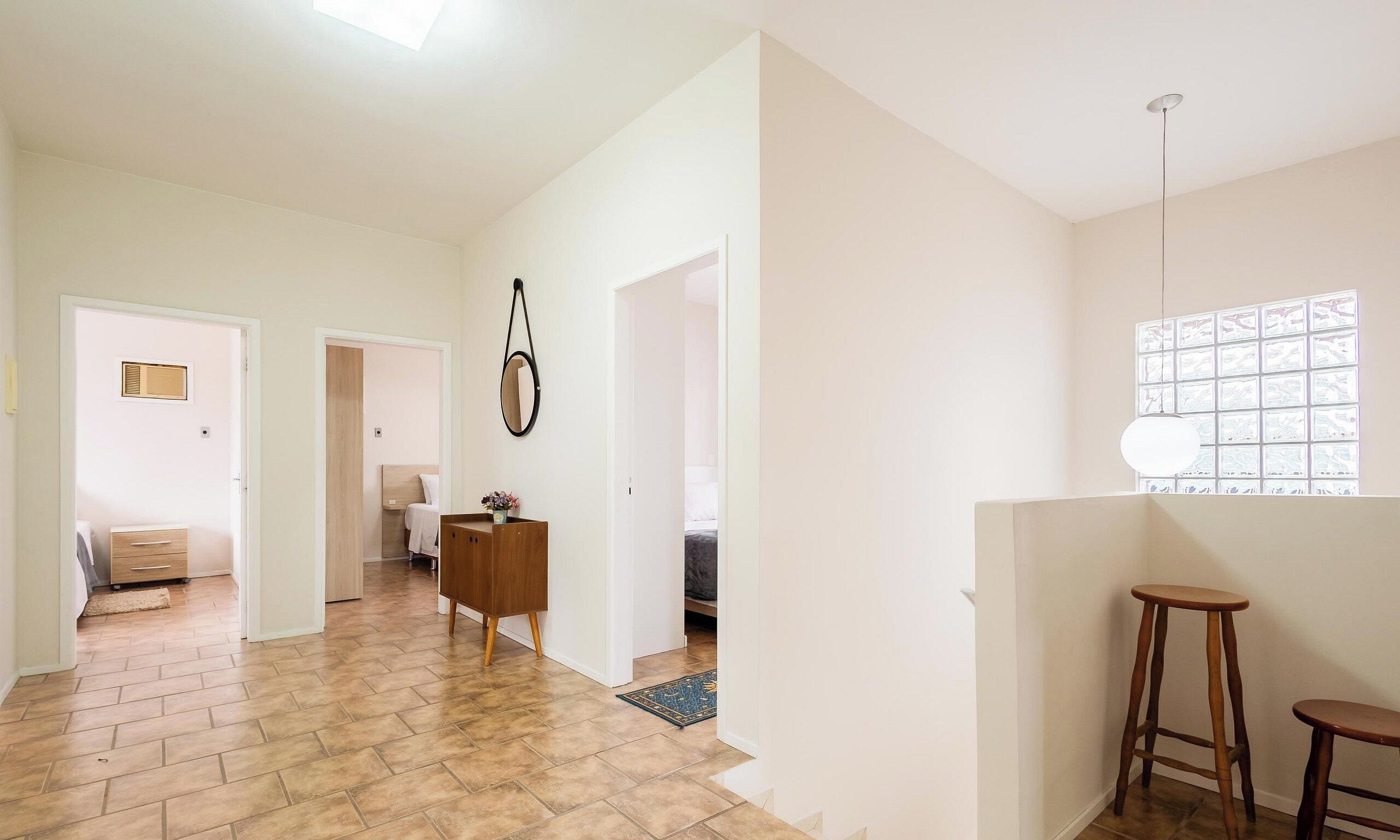 Un pasillo amplio con suelo de madera, paredes blancas y un mueble de madera con un espejo. A la derecha, hay una ventana con persianas.