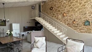 Uma área de refeições com uma mesa de madeira e cadeiras de metal, uma cozinha com geladeira e fogão, e uma escadaria com parede de pedra.