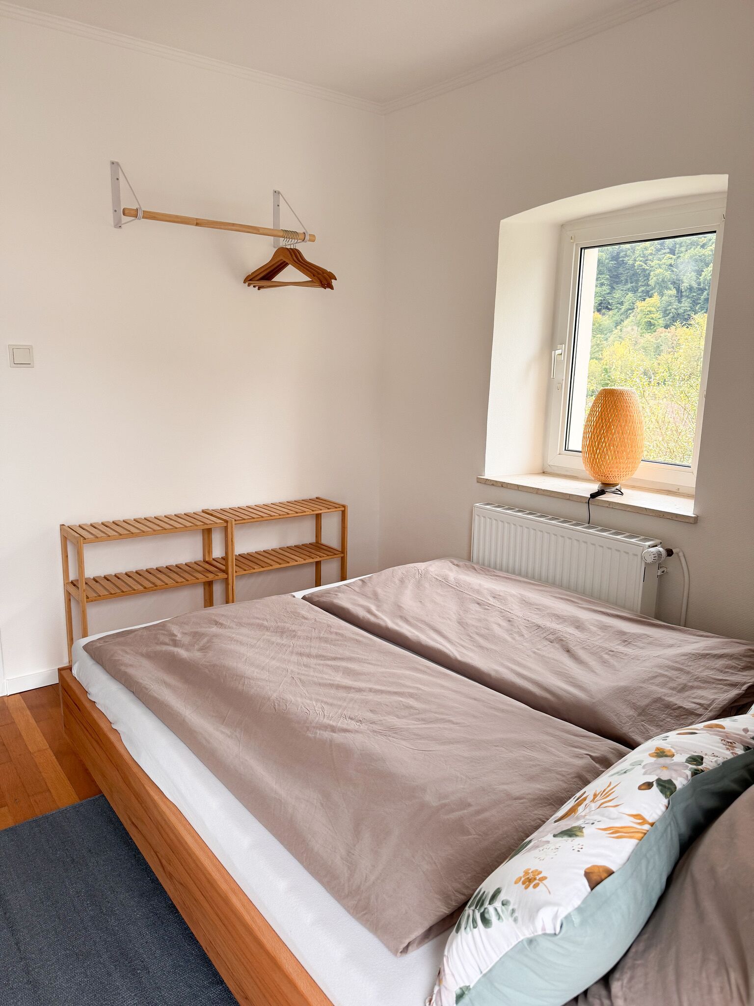 Een slaapkamer met een bed, een radiator, een raam met uitzicht op groen en een houten plank.