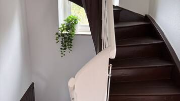 Una escalera de madera con balaustres blancos y barandilla, que lleva a un rellano con una ventana y una planta colgante.