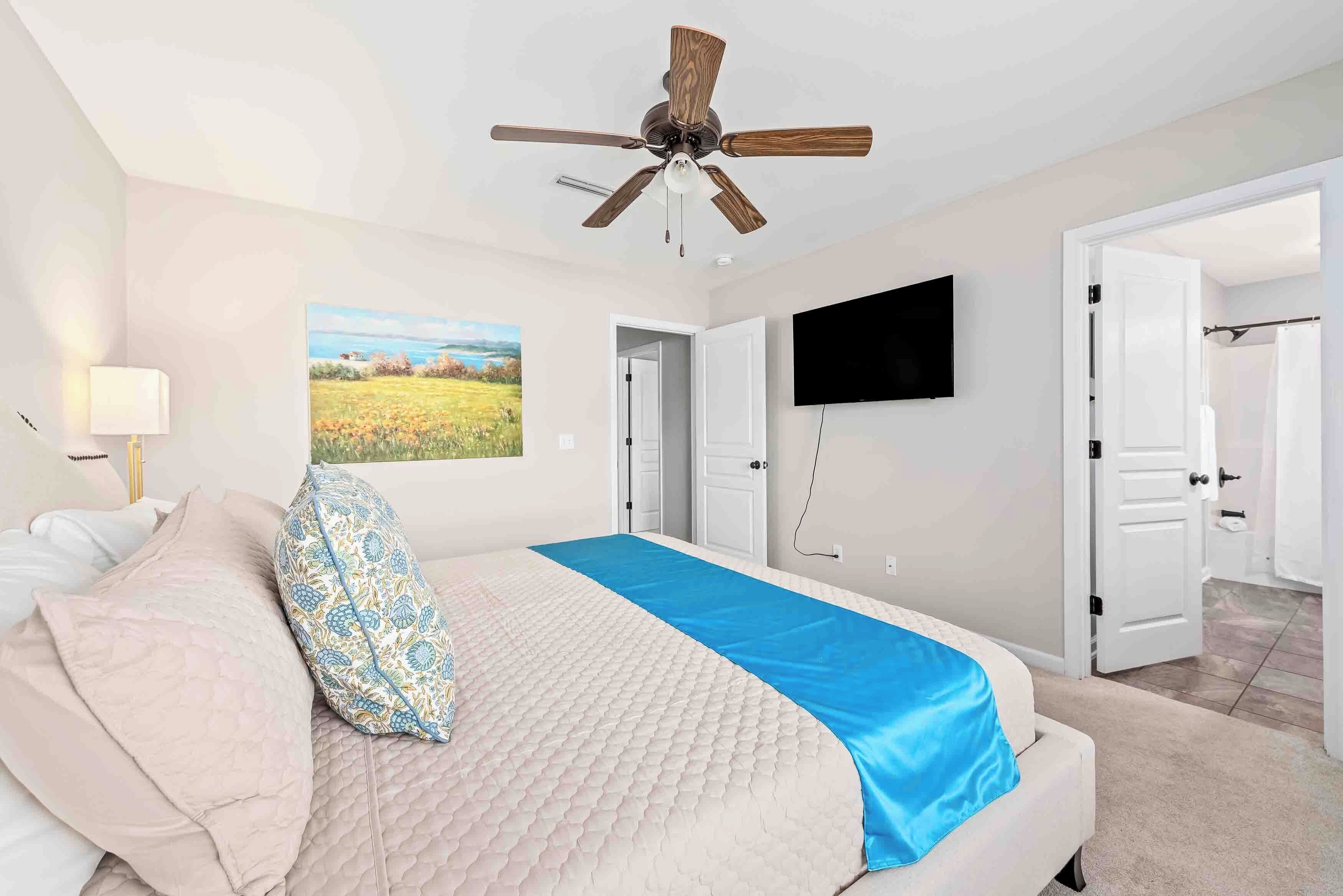 A bedroom with a bed, a ceiling fan, a TV mounted on the wall, and a door leading to another room.