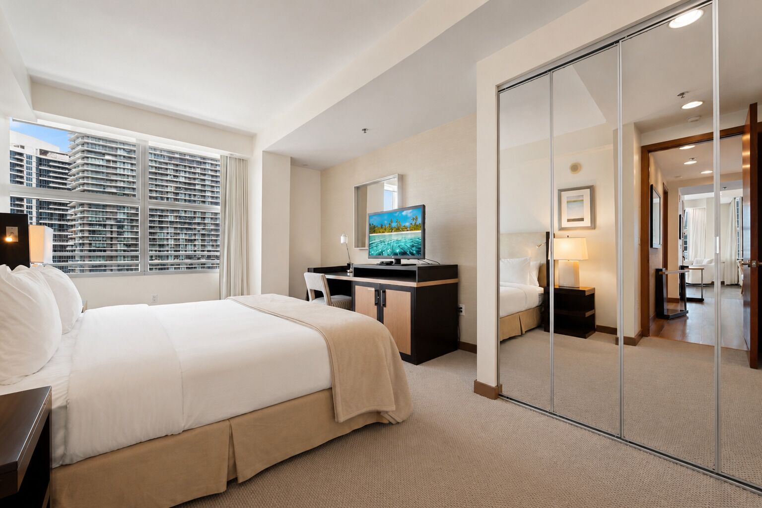 A hotel room with a large bed, a desk with a TV, a mirror, and a view of high-rise buildings.