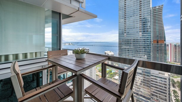 A balcony with a table and chairs, a potted plant, and a view of a tall building and a bridge. - Brickell Sea View 2BR Free Parking by Palermo Home (Miami)