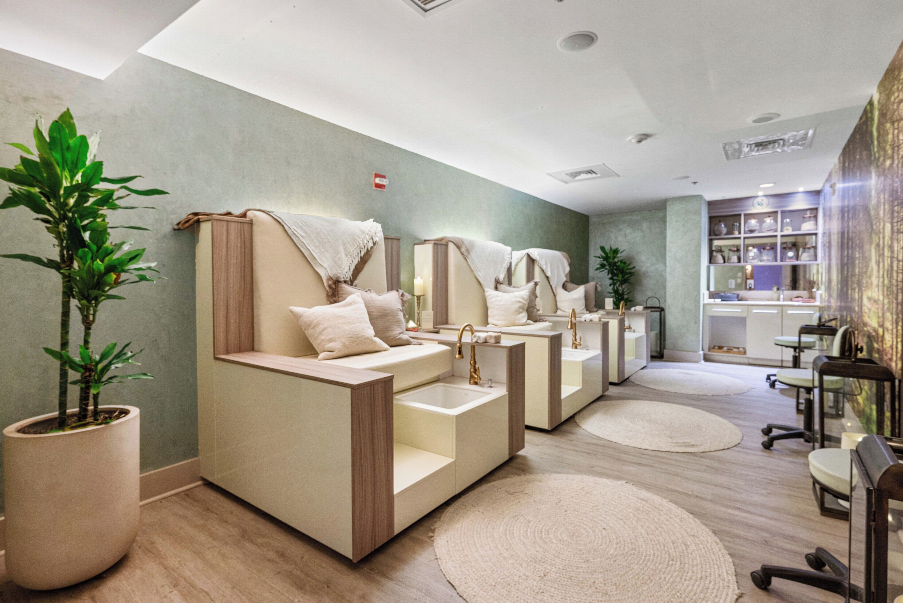 Une salle de spa dotée de plusieurs postes de soins, chacun comprenant un banc rembourré, un lavabo et un miroir. Des plantes en pot et un grand miroir ornent le mur.