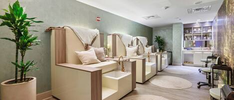 Une salle de spa dotée de plusieurs postes de soins, chacun comprenant un banc rembourré, un lavabo et un miroir. Des plantes en pot et un grand miroir ornent le mur.