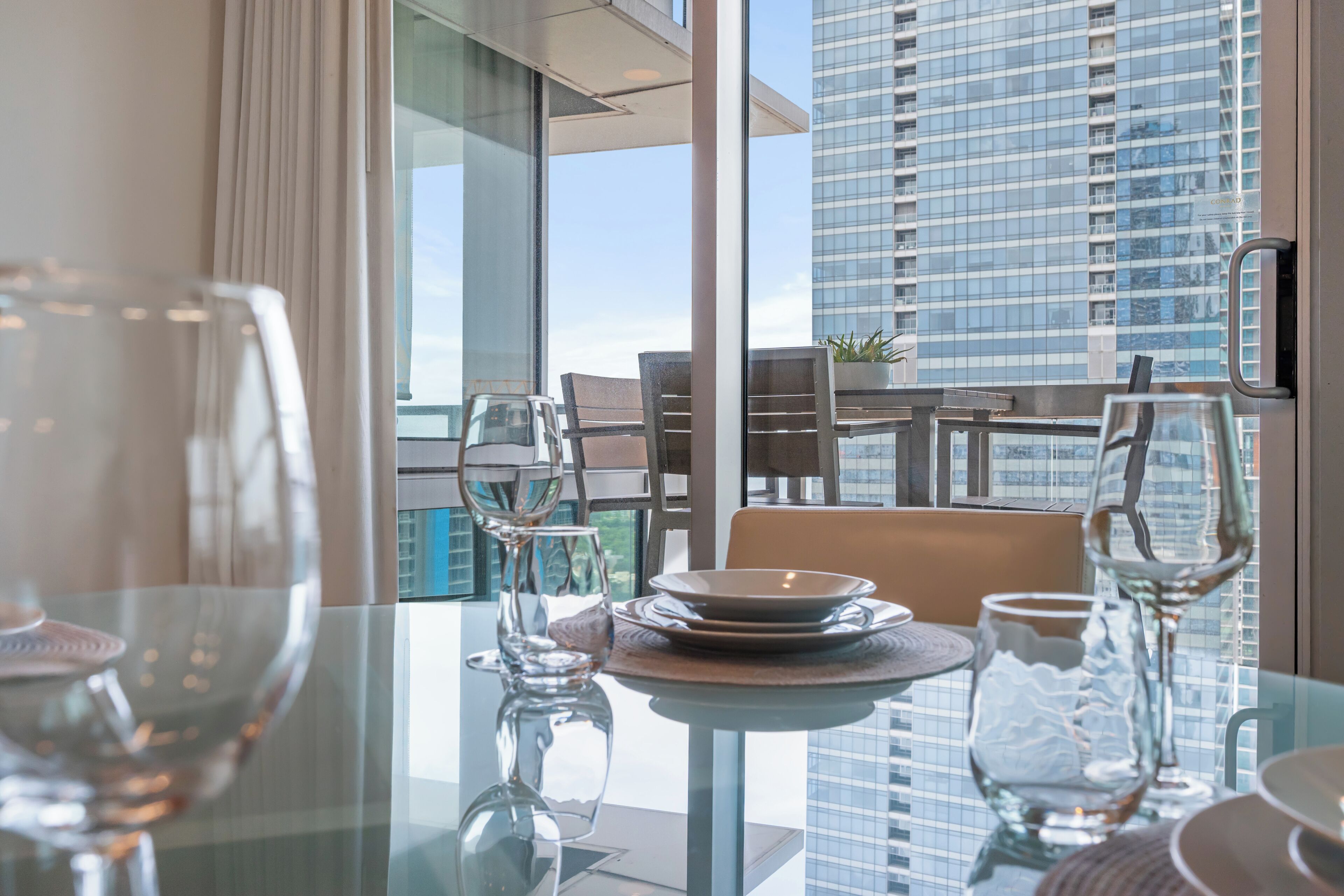 Une table à manger avec de la verrerie et des assiettes, une vue sur un immeuble de grande hauteur et un balcon avec des chaises et une table.