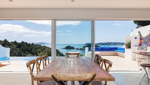 A wooden dining table with chairs, a blue sofa, and a view of the sea through large glass doors. - Seabreeze Sanctuary - Paihia Holiday Home (Paihia)