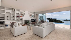 A spacious living room with a large window overlooking a scenic view. There are two sofas, a glass coffee table, and a fireplace. - Seabreeze Sanctuary - Paihia Holiday Home (Paihia)
