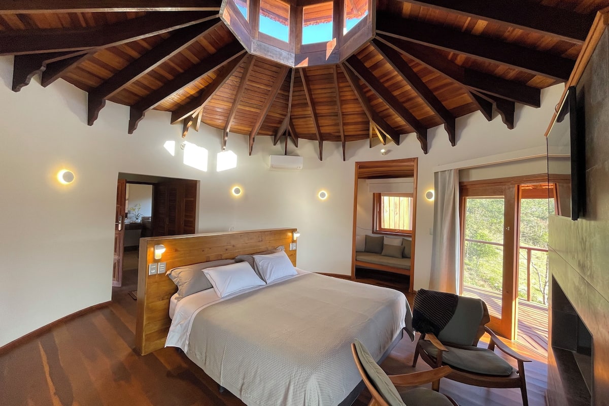 A bedroom with a wooden ceiling, a bed, a chair, and a window with a view of trees.