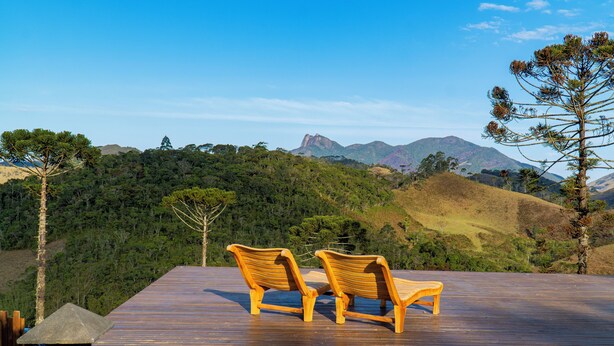 A wooden deck with two wooden chairs facing a scenic view of a forested mountain range. - Atiaia Jungle River Retreat (Bocaina de Minas)