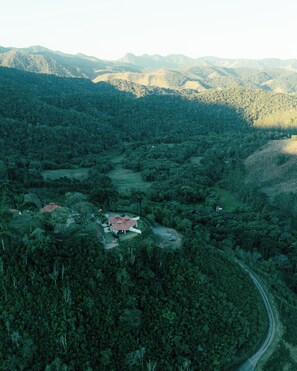 Vue aérienne d’une zone boisée avec un petit groupe de bâtiments, dont une maison avec un toit rouge et une construction plus importante avec un toit blanc, le tout entouré d’arbres denses et de collines en arrière-plan.
