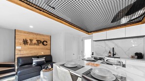 A modern living room with a black leather sofa, a wooden accent wall with #LPC, a dining area with a white table set for two, and a kitchen with white cabinets and a marble backsplash. - Luxury mini-chalets to disconnect for two with a private spa nestled in the hear (VAL DES LACS)