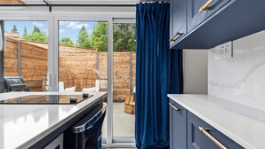 A modern kitchen with a large window, a sliding glass door leading to an outdoor seating area, and a view of a wooden deck and trees. - Luxury mini-chalets to disconnect for two with a private spa nestled in the hear (VAL DES LACS)