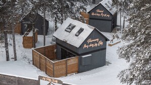 Una cabaña rústica con una terraza de madera y un pequeño porche, rodeada de árboles cubiertos de nieve.