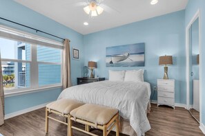 A bedroom with a large bed, a window with blinds, a painting of a boat, and a nightstand with lamps. - Baywatch H3 (Pensacola Beach)