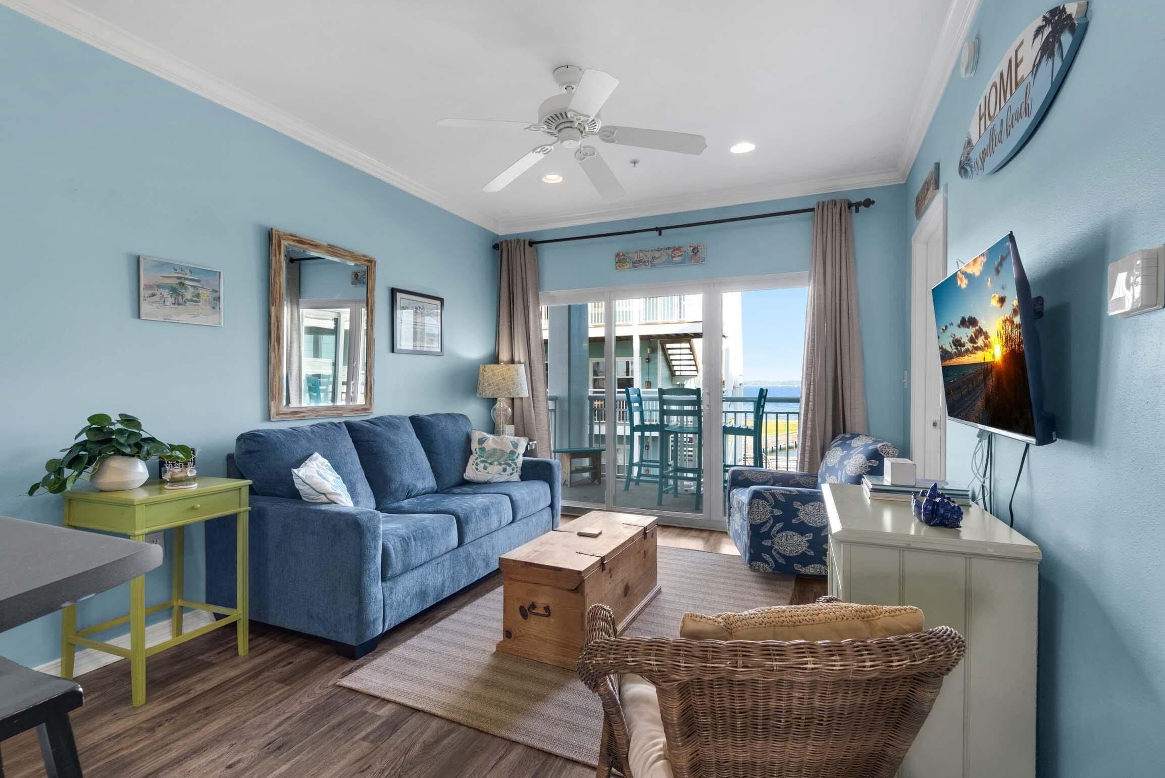 A living room with a blue sofa, a wooden chest, a wicker chair, and a television on the wall.